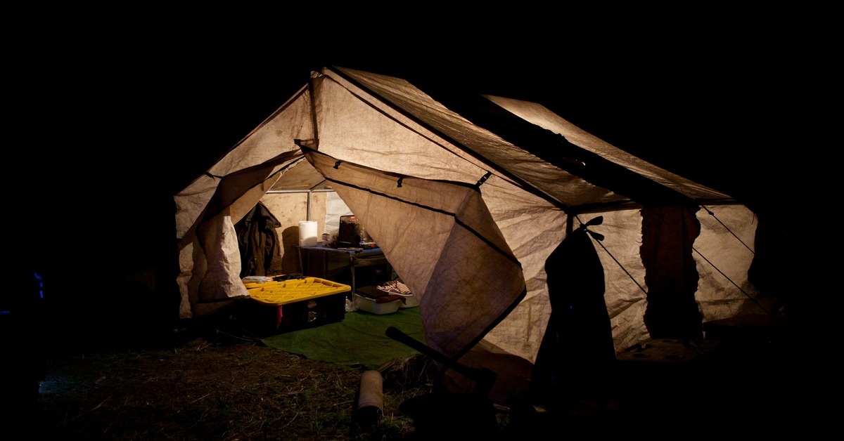 An illuminated tent at night. Through the opening you can see a storage tub, a table, and various supplies.